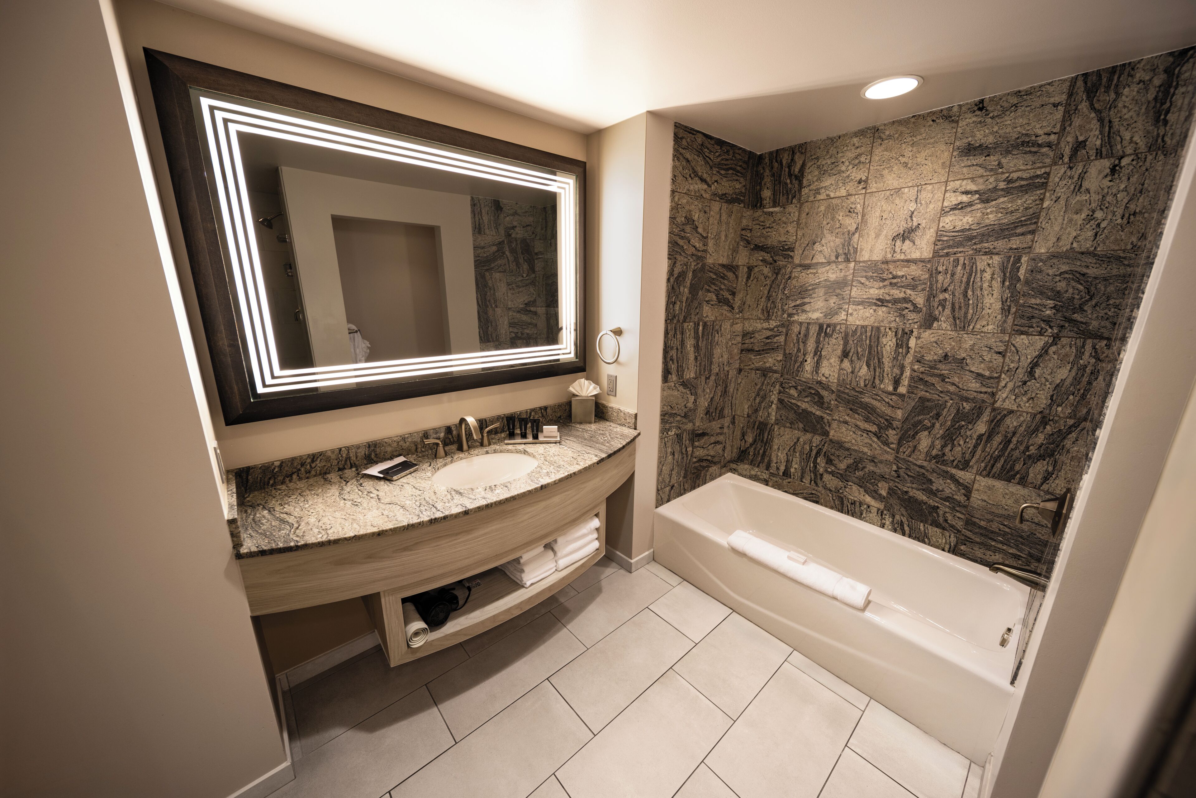 A modern bathroom with a large mirror, two sinks, a bathtub, and a stone-textured wall.