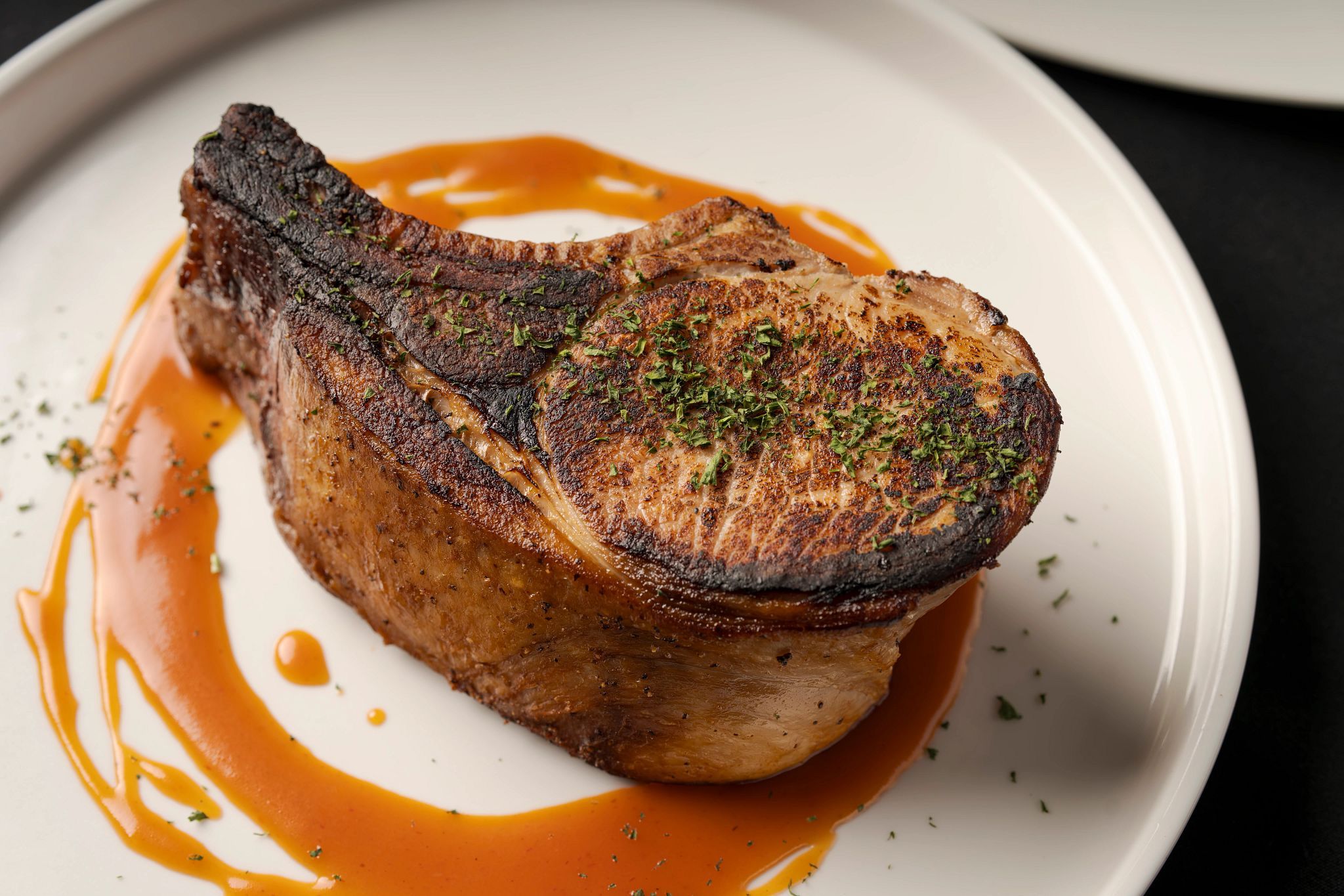 A steak with a charred crust, garnished with herbs, on a white plate with a sauce.