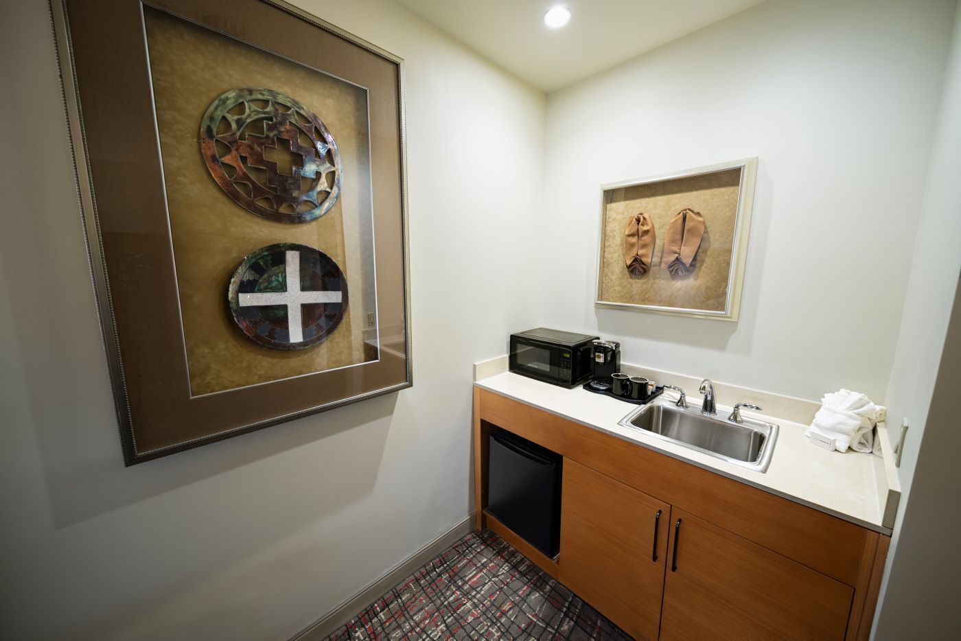 Une chambre d’hôtel de taille réduite, dotée d’une kitchenette, d’un évier et de deux œuvres d’art encadrées au mur.