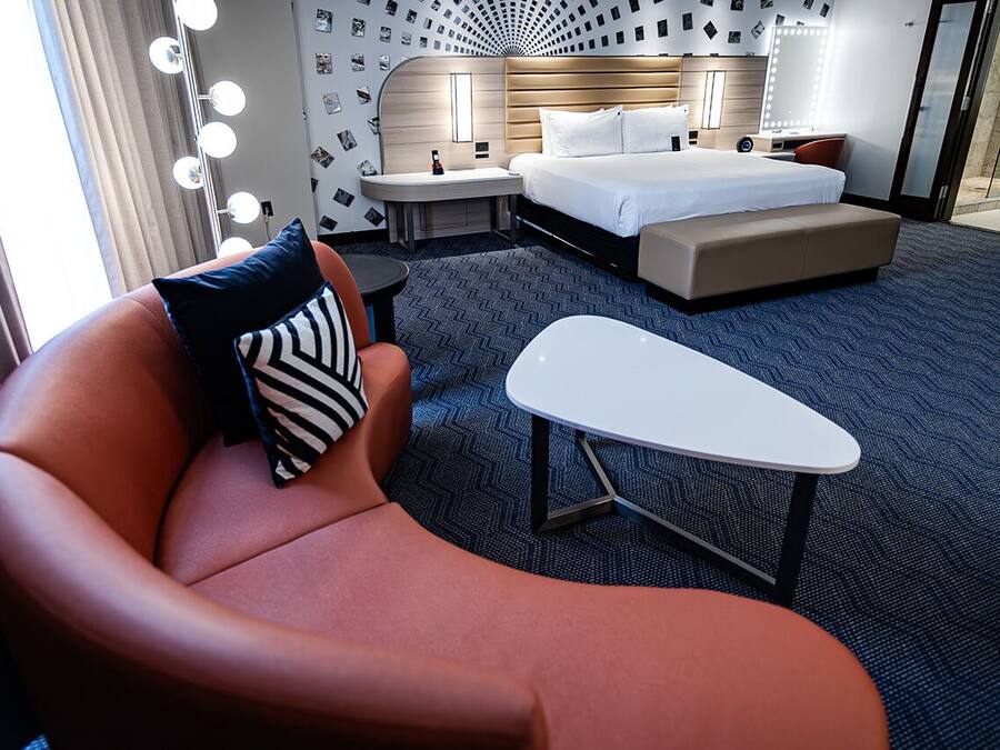 A hotel room with a curved orange chair, a small white table, a bed with a headboard, and a wall with a patterned wallpaper.