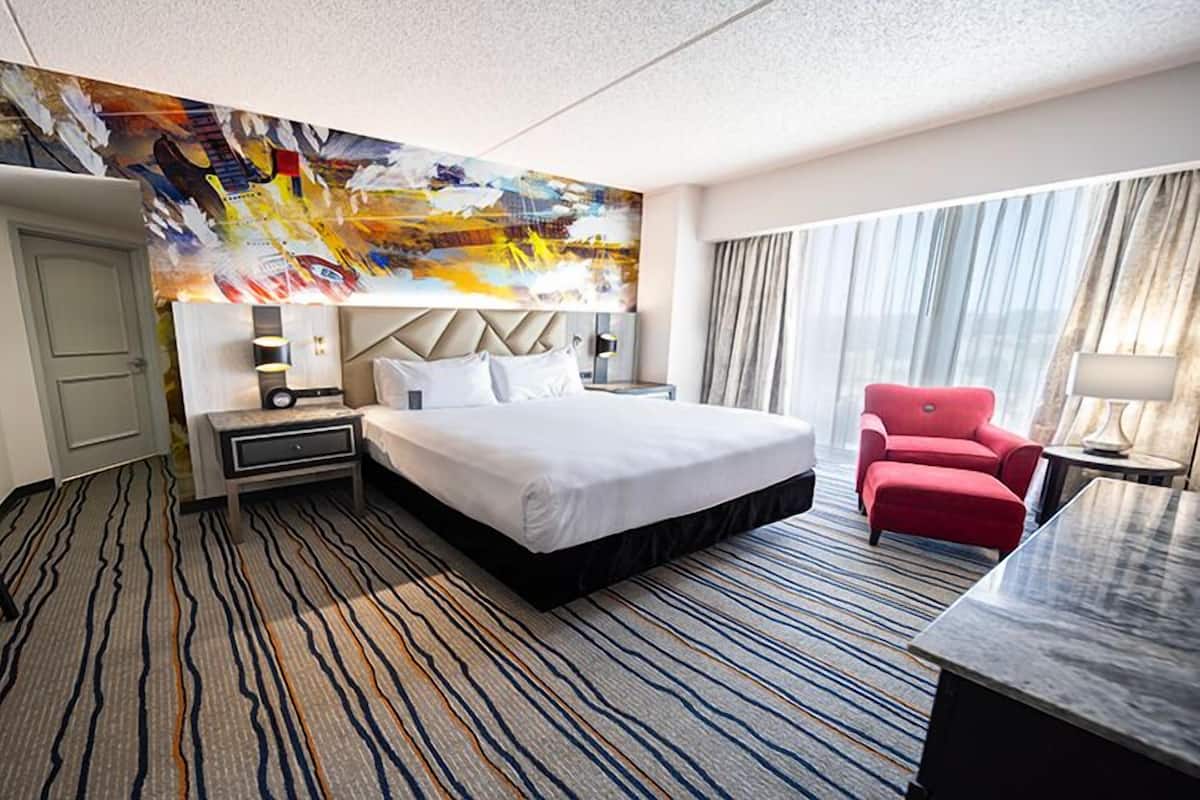 Habitación de hotel con cama, escritorio, silla, sillón rojo, un cuadro en la pared y alfombra estampada.