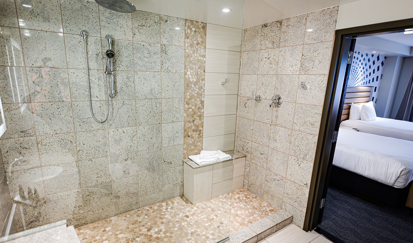 A hotel bathroom with a shower, a toilet, and a sink. There is a bed visible through an open door.