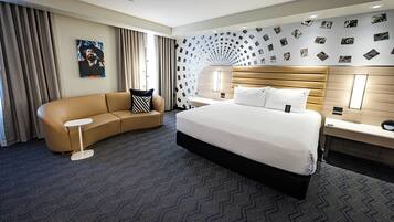 A modern hotel room with a large bed, a curved sofa, a small side table, and a wall decorated with photographs.