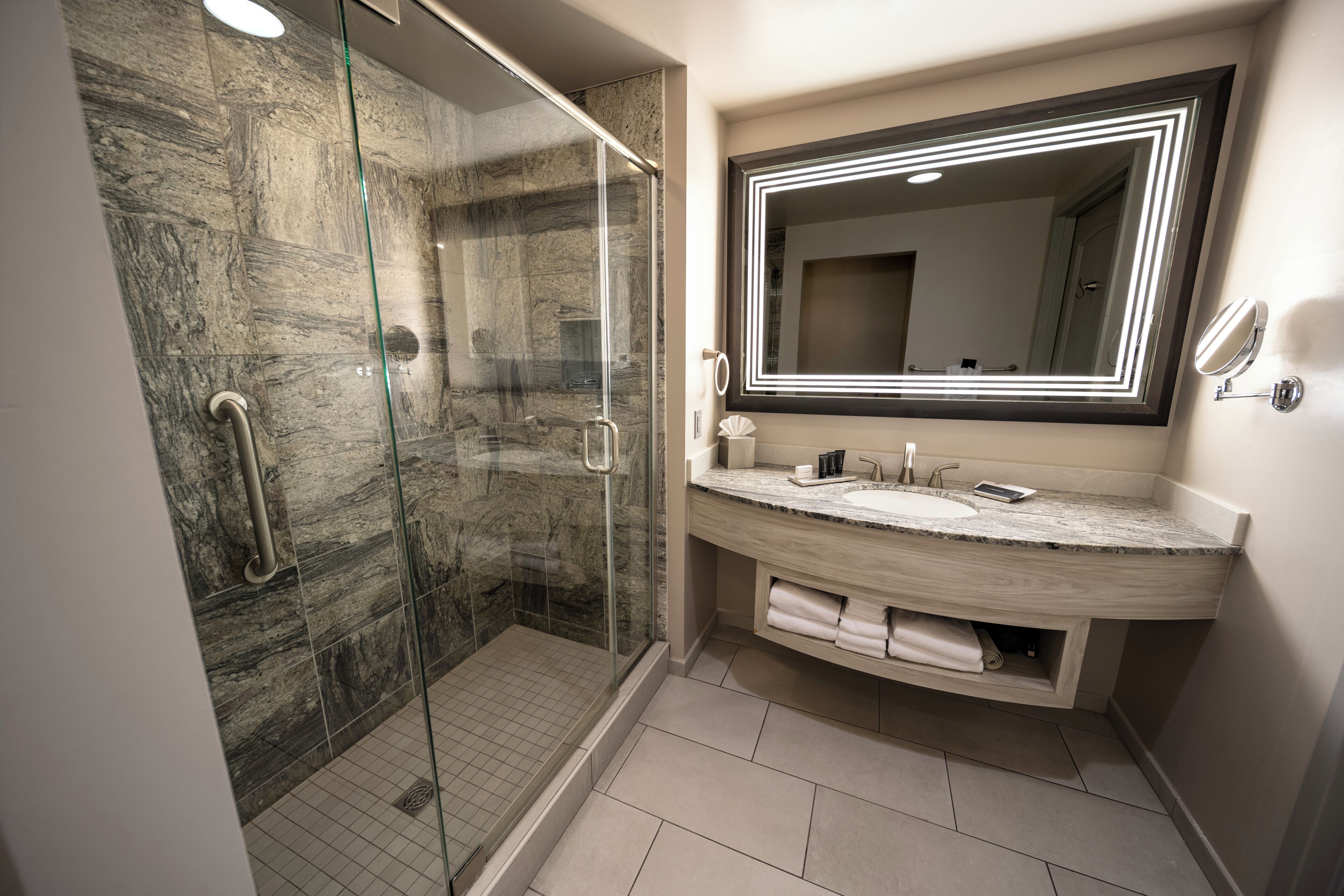 Une salle de bain moderne dotée d’une douche à parois de verre, d’un mur recouvert de carreaux de marbre, d’un grand miroir avec éclairage intégré et d’un meuble-lavabo en bois.