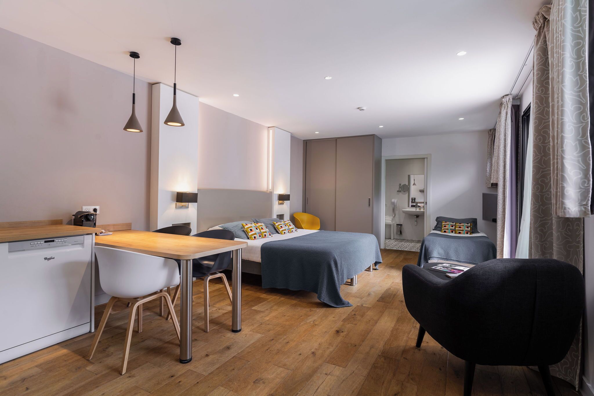 Ein modernes, kleines Schlafzimmer mit Holzboden, einem Bett mit blauer Bettdecke, einem Schreibtisch mit Geschirrspüler und einem Stuhl.