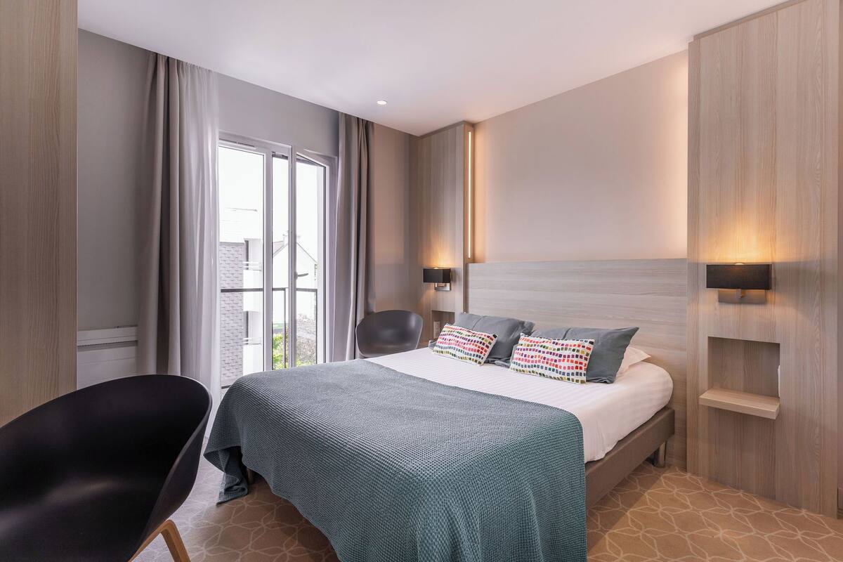 A hotel room with a bed, a chair, a window with curtains, and wall-mounted lighting.