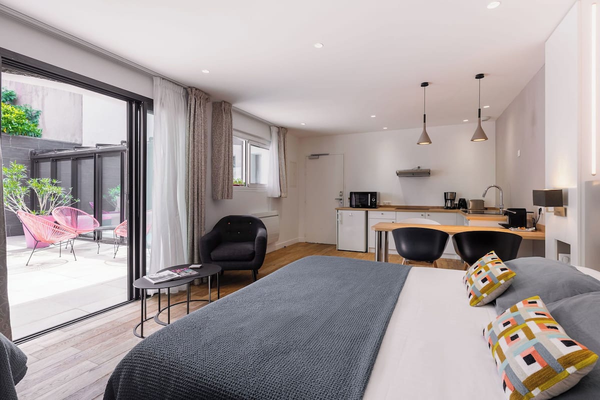 A modern hotel room with a large bed, a desk, and a view of a balcony with chairs.