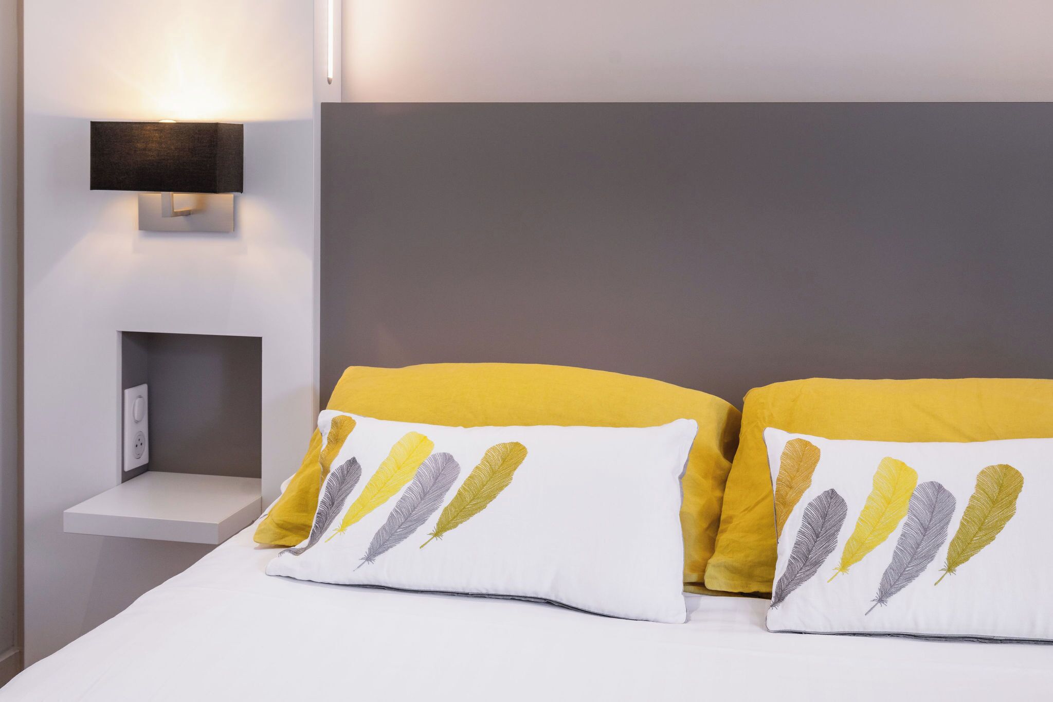 A neatly made bed with white and yellow bedding, a gray headboard, and a wall-mounted light fixture.