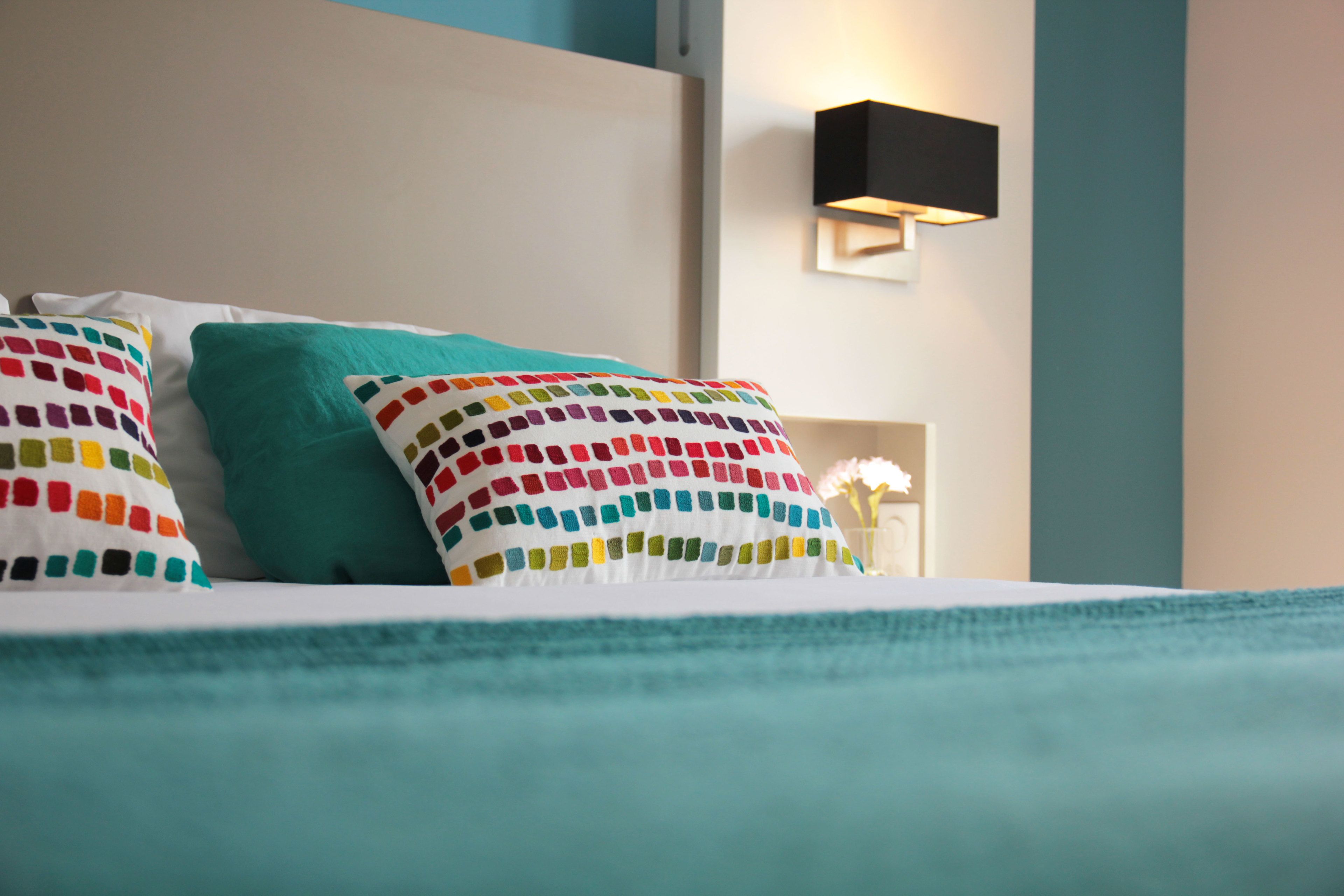 Una cama con ropa de cama color turquesa y almohadas coloridas, una lámpara fijada a la pared y un pequeño florero con flores.