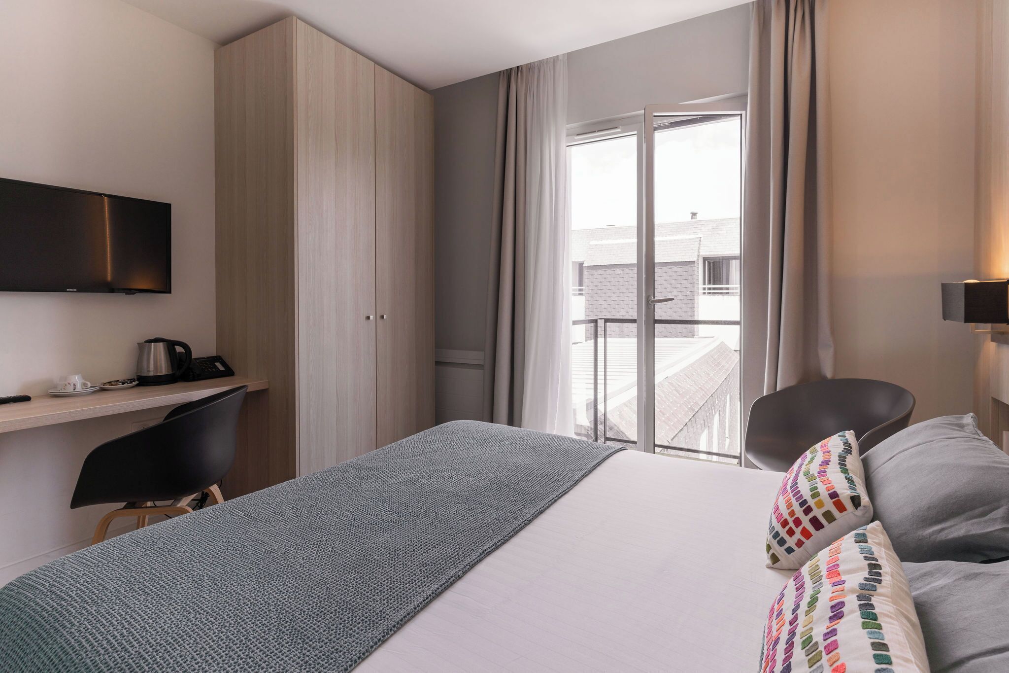 Una habitación de hotel con cama, escritorio, silla, televisor y balcón con cortinas.