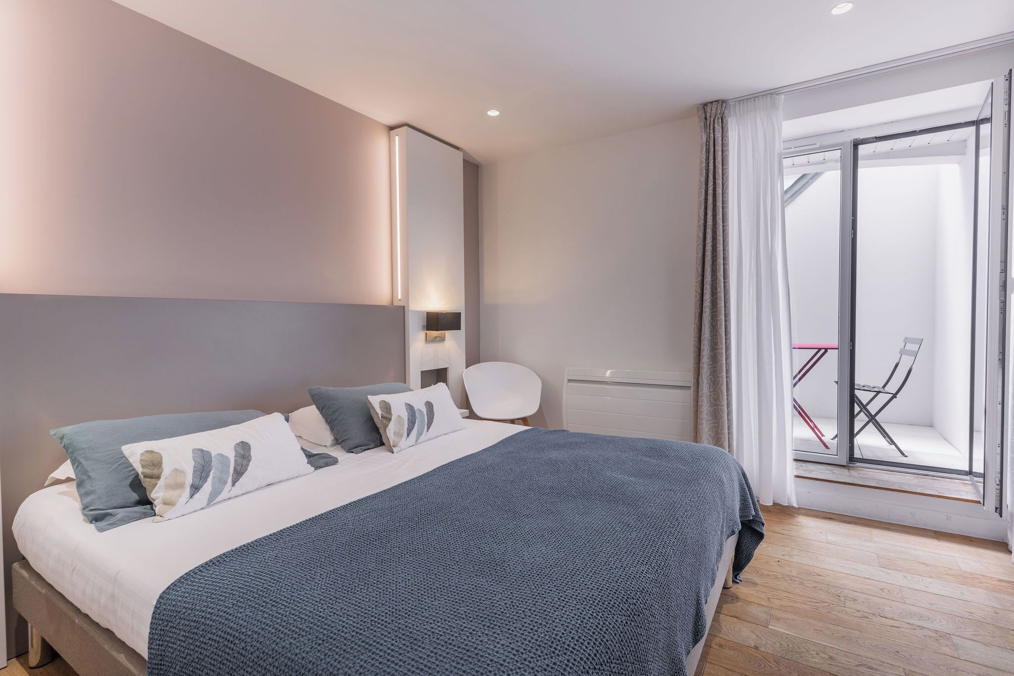 A modern bedroom with a large bed, a bedside table, a chair, and a sliding glass door leading to a balcony.