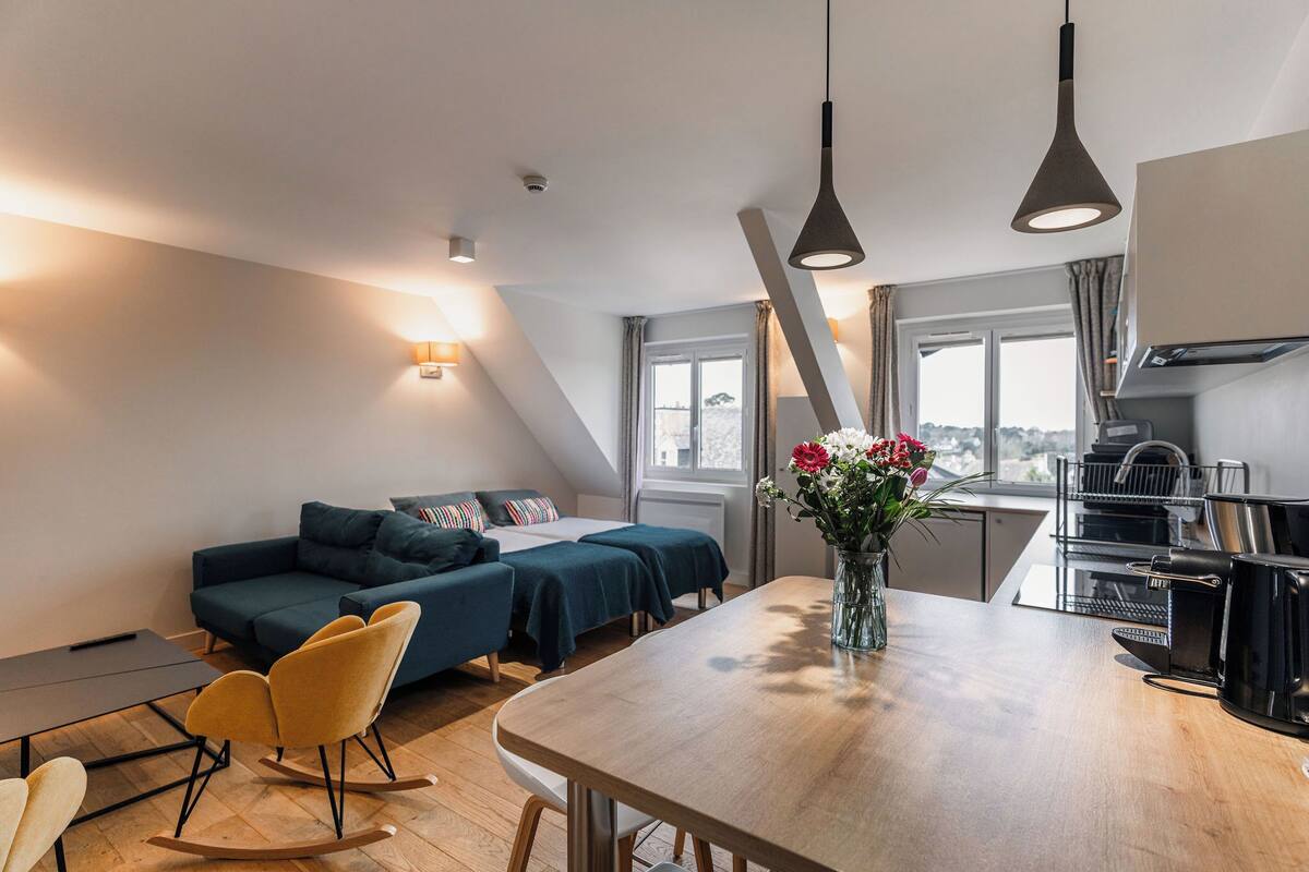 A modern room with a sofa, two beds, a dining table, and a kitchen area.