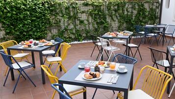 Un área de comedor al aire libre con mesas y sillas, rodeada por un muro con follaje.