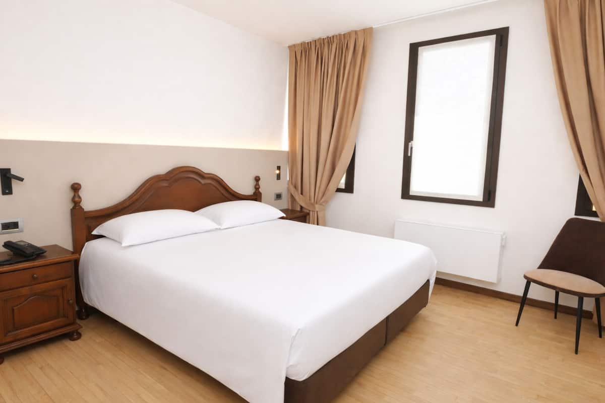 A neatly made bed in a hotel room with a wooden headboard, a bedside table with a phone, a chair, and a window with curtains.