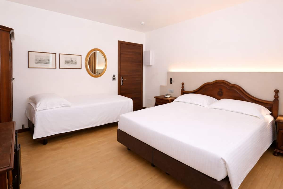 A hotel room with a large bed, a smaller bed, a wooden wardrobe, a bedside table with a lamp, and two framed pictures on the wall.