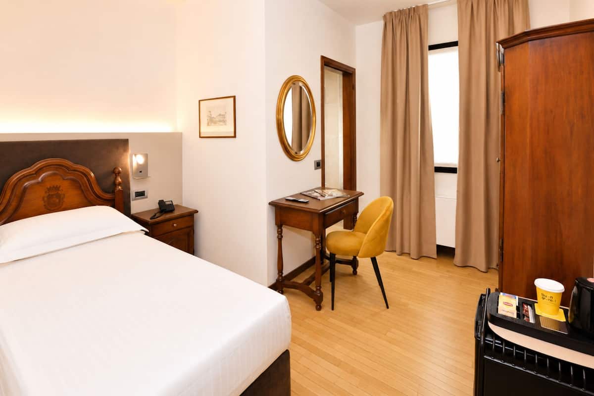 A hotel room with a bed, a desk, a chair, a mirror, a wardrobe, and a coffee machine.