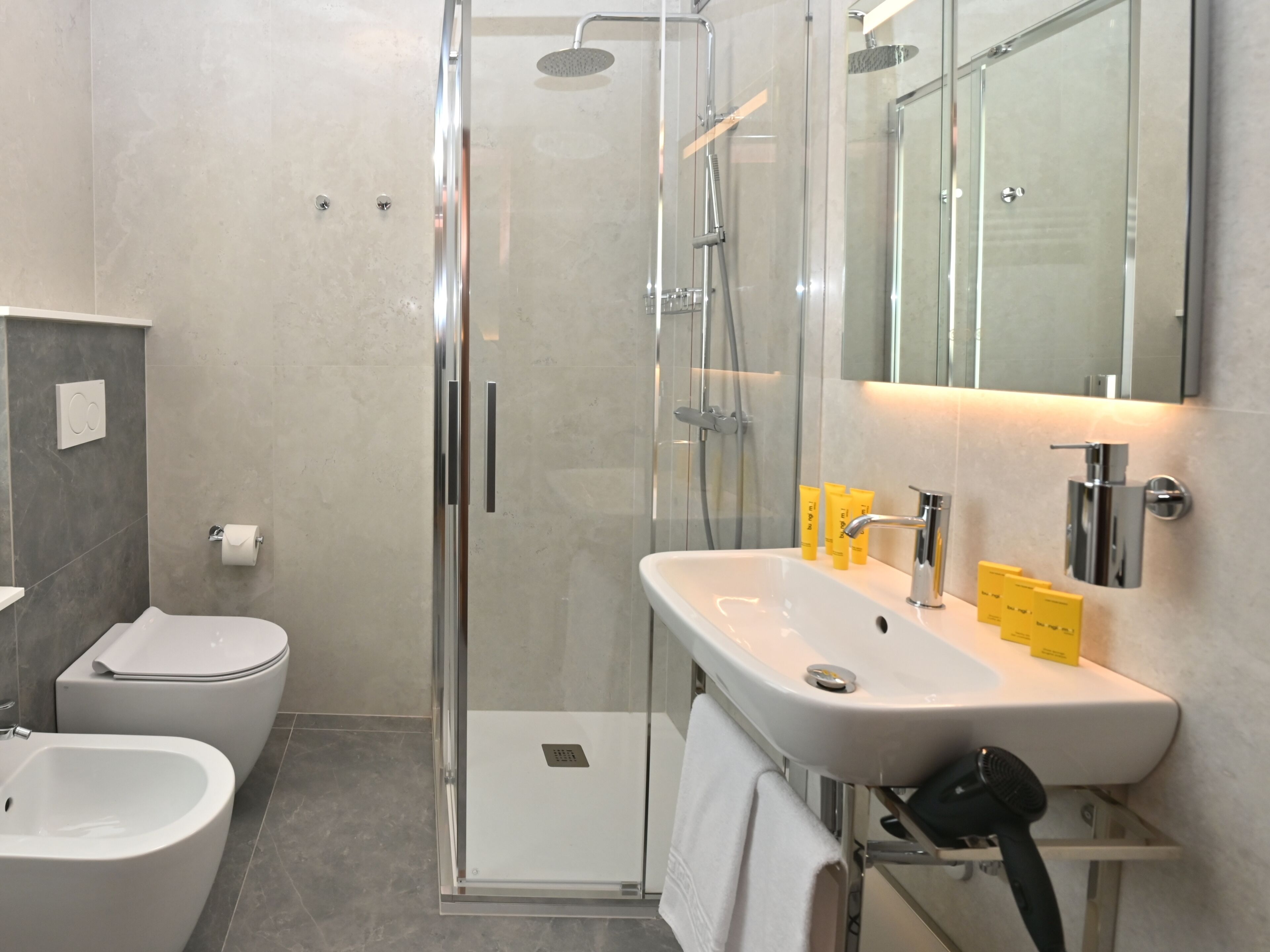 A modern bathroom with a glass-enclosed shower, a white sink with yellow soap, a toilet, and a bidet.