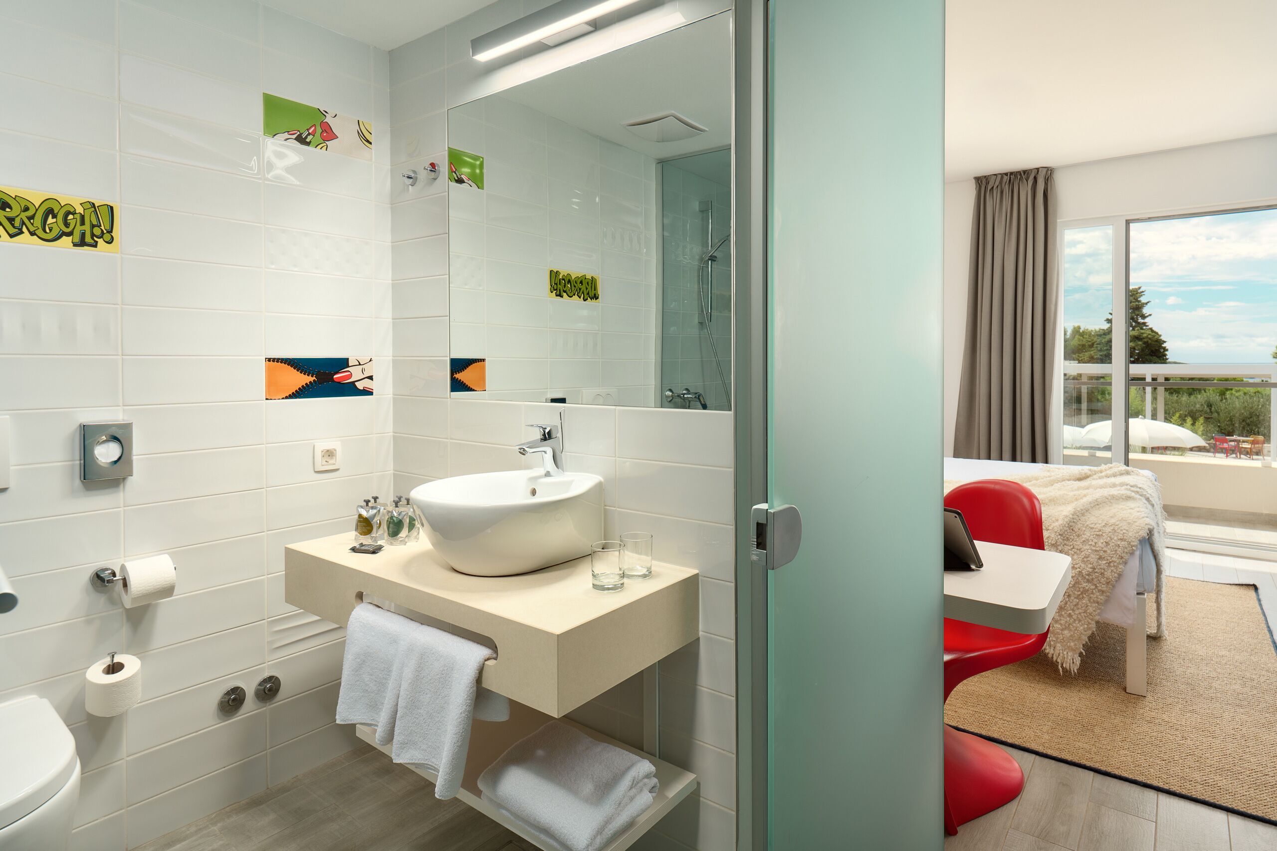 Un baño moderno con un espejo grande, un lavamanos y un inodoro. Cuenta con una puerta de ducha de vidrio y una ventana pequeña con cortinas.