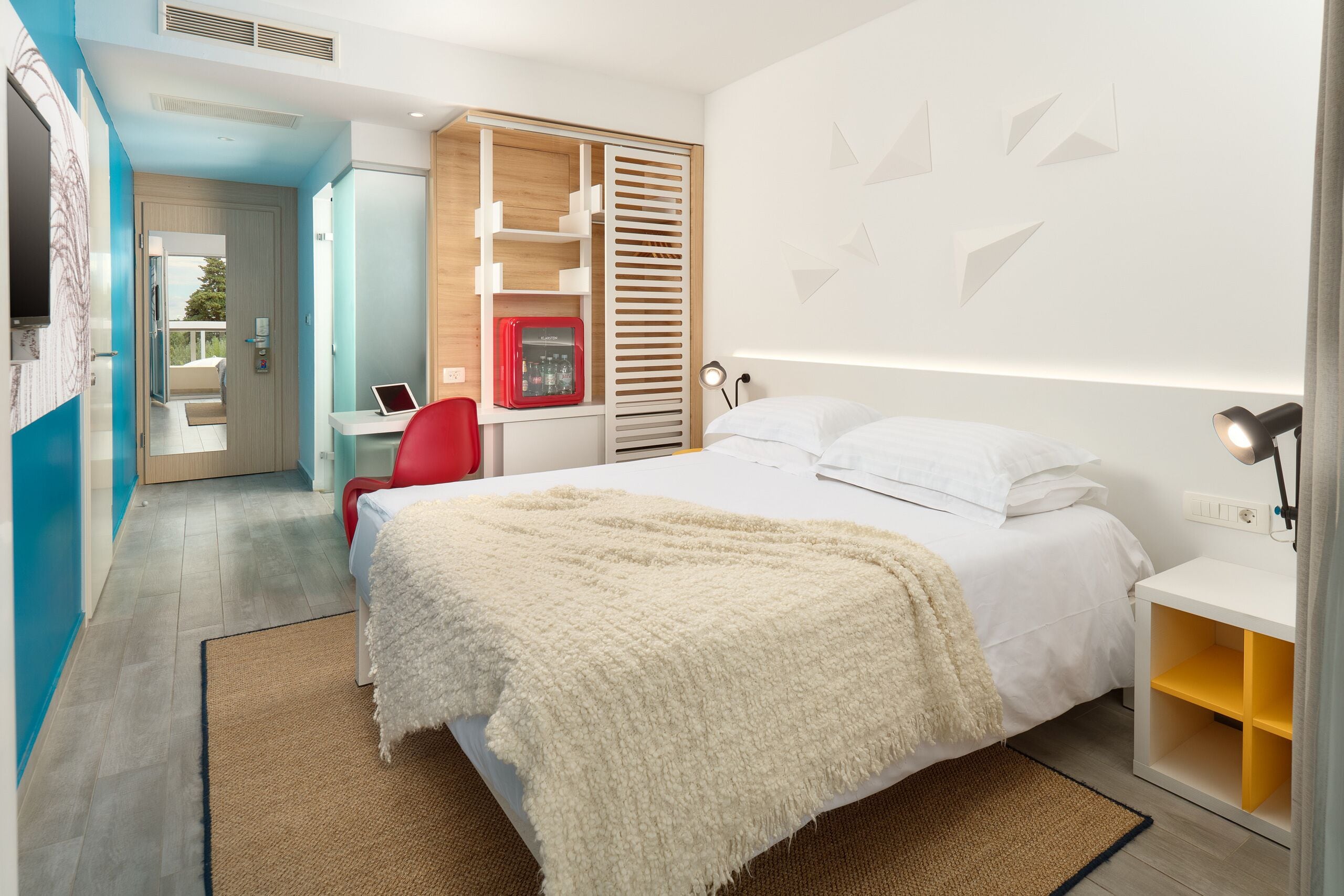 A modern hotel room with a bed, bedside table, and a desk with a computer.