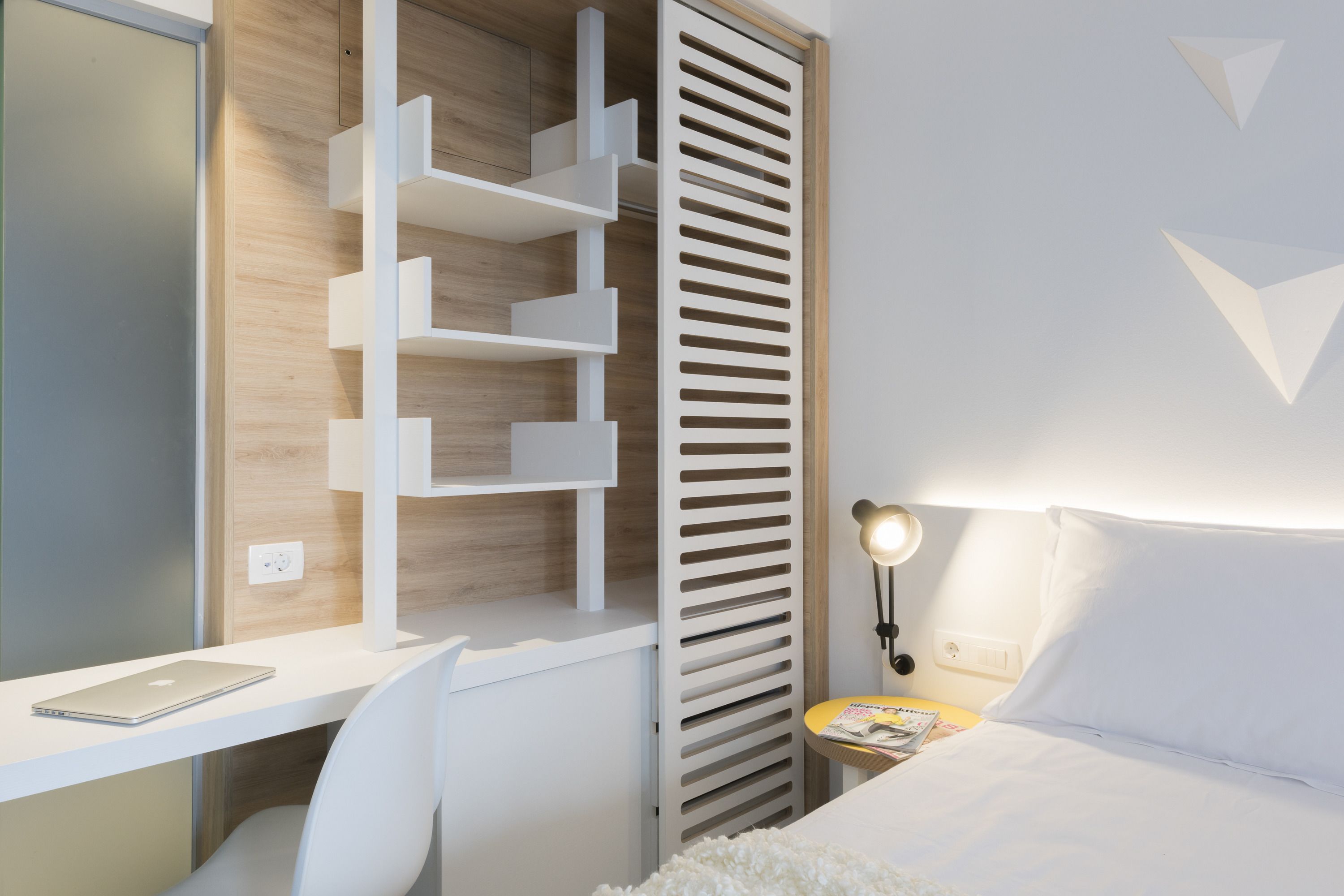 A modern bedroom with a white desk, a chair, a bed with white bedding, a bedside table with a lamp, and a built-in shelving unit.