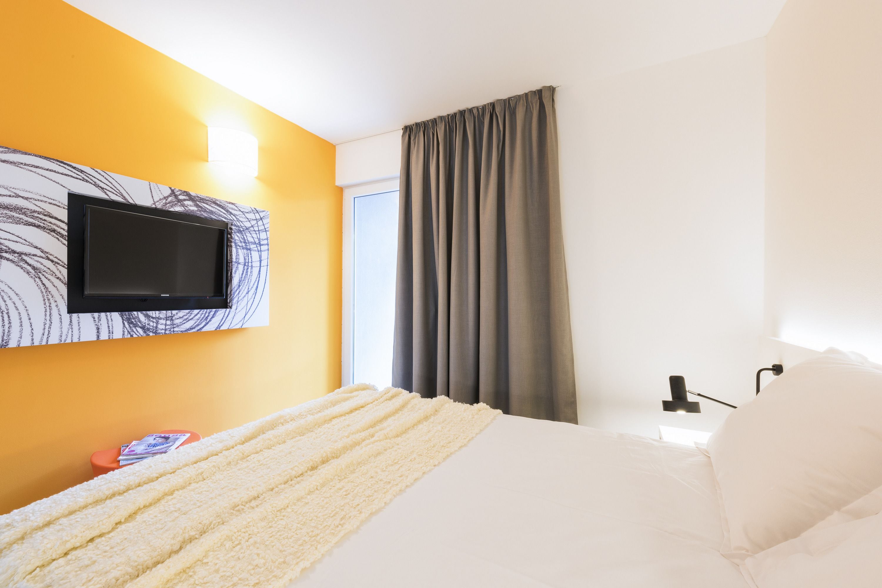 Habitación de hotel con cama, televisor de pantalla plana montado en la pared, ventana con cortinas y mesita de noche con lámpara.