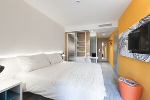 Una habitación de hotel moderna con una cama, un escritorio, una silla, un televisor y un baño visible a través de una puerta abierta.