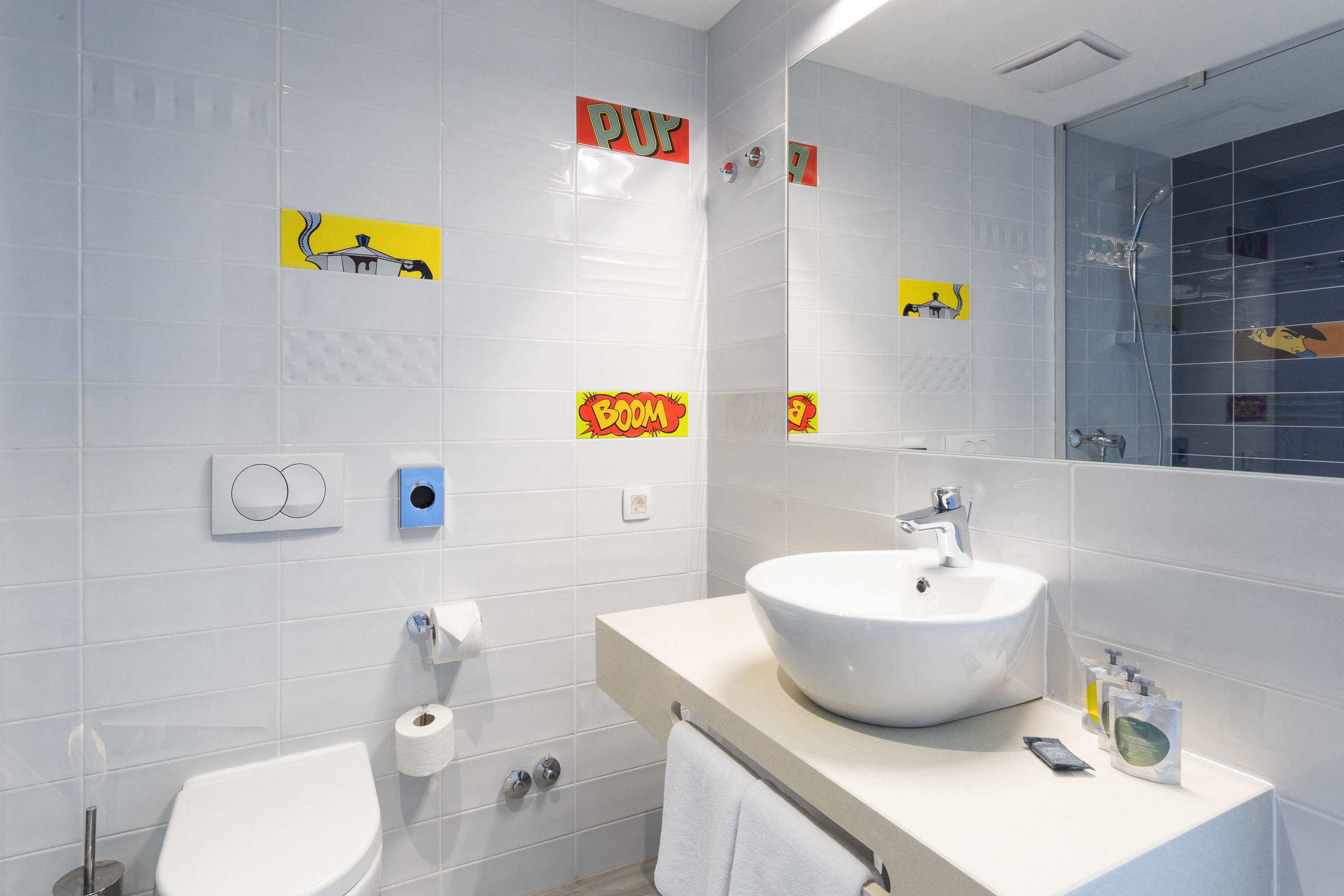 A modern bathroom with a white sink, toilet, and shower. There are stickers on the wall and a mirror above the sink.