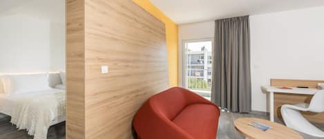 A modern hotel room with a wooden accent wall, a red sofa, a small round table, and a bed with white bedding.