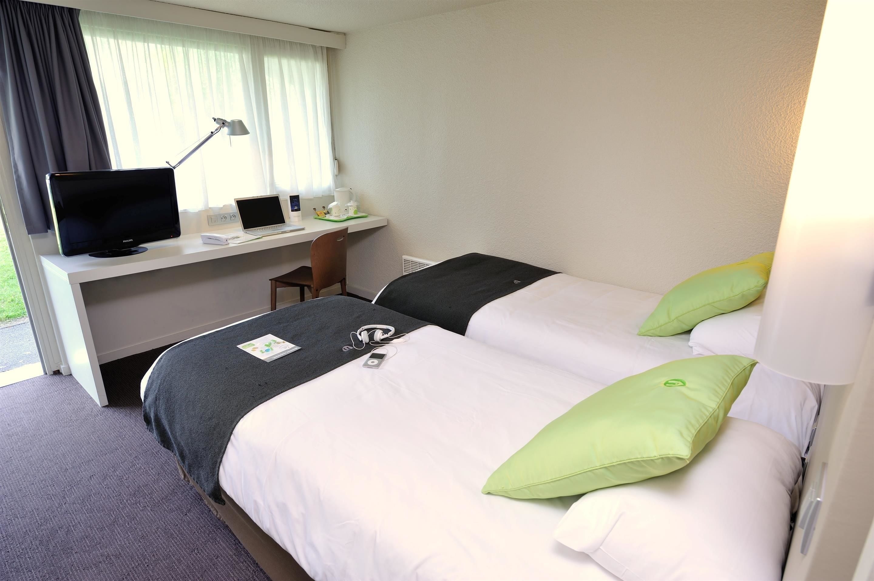 Habitación de hotel con dos camas, un escritorio con computadora y lámpara, y una ventana con cortinas.