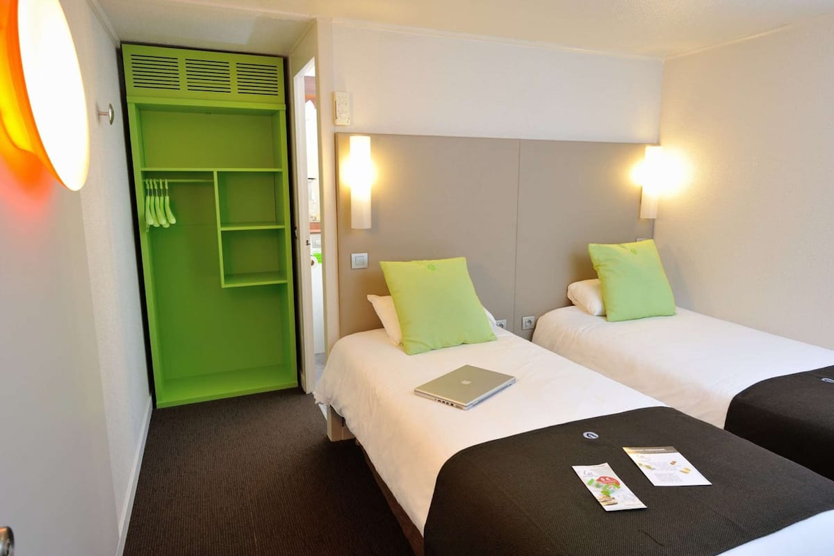 Ein Hotelzimmer mit zwei Betten, jeweils mit einem Kissen und einem Laptop. Es gibt einen grünen Kleiderschrank und eine wandmontierte Leuchte.