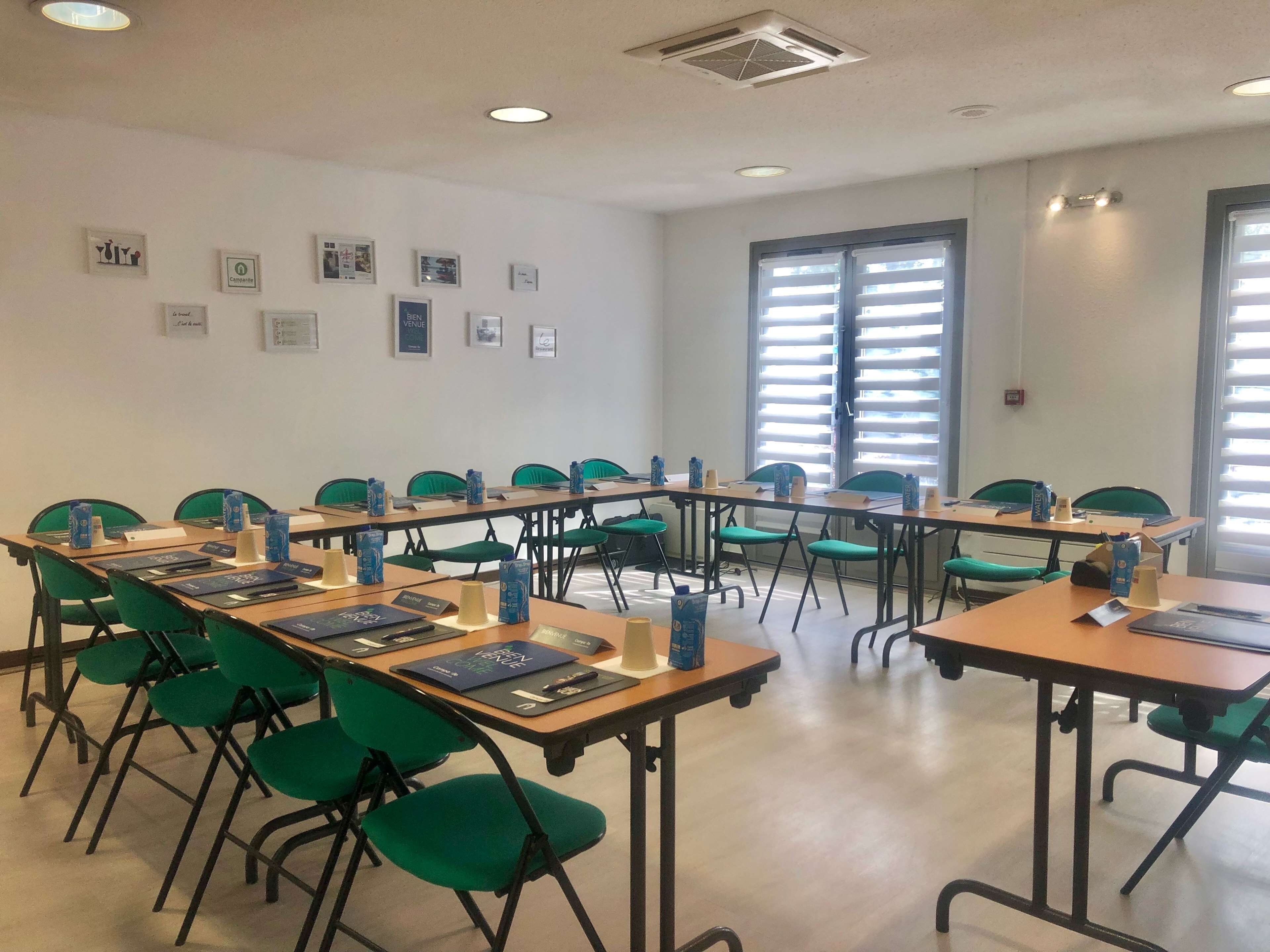 Une salle de conférence avec des chaises et des tables vertes, des tableaux encadrés aux murs et une fenêtre avec des stores.