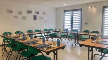 Une salle de conférence avec des chaises et des tables vertes, des tableaux encadrés aux murs et une fenêtre avec des stores.