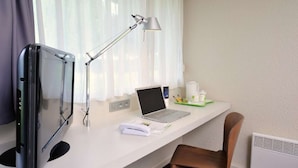 Een hotelkamer met een bureau, een laptop, een bureaulamp, een flatscreen televisie, een stoel en een koffiekop.