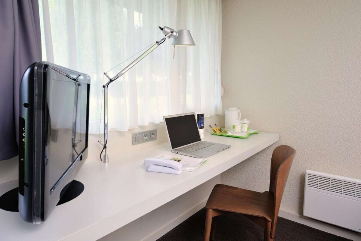 A hotel room with a desk, a laptop, a desk lamp, a flat-screen TV, a chair, and a coffee cup.