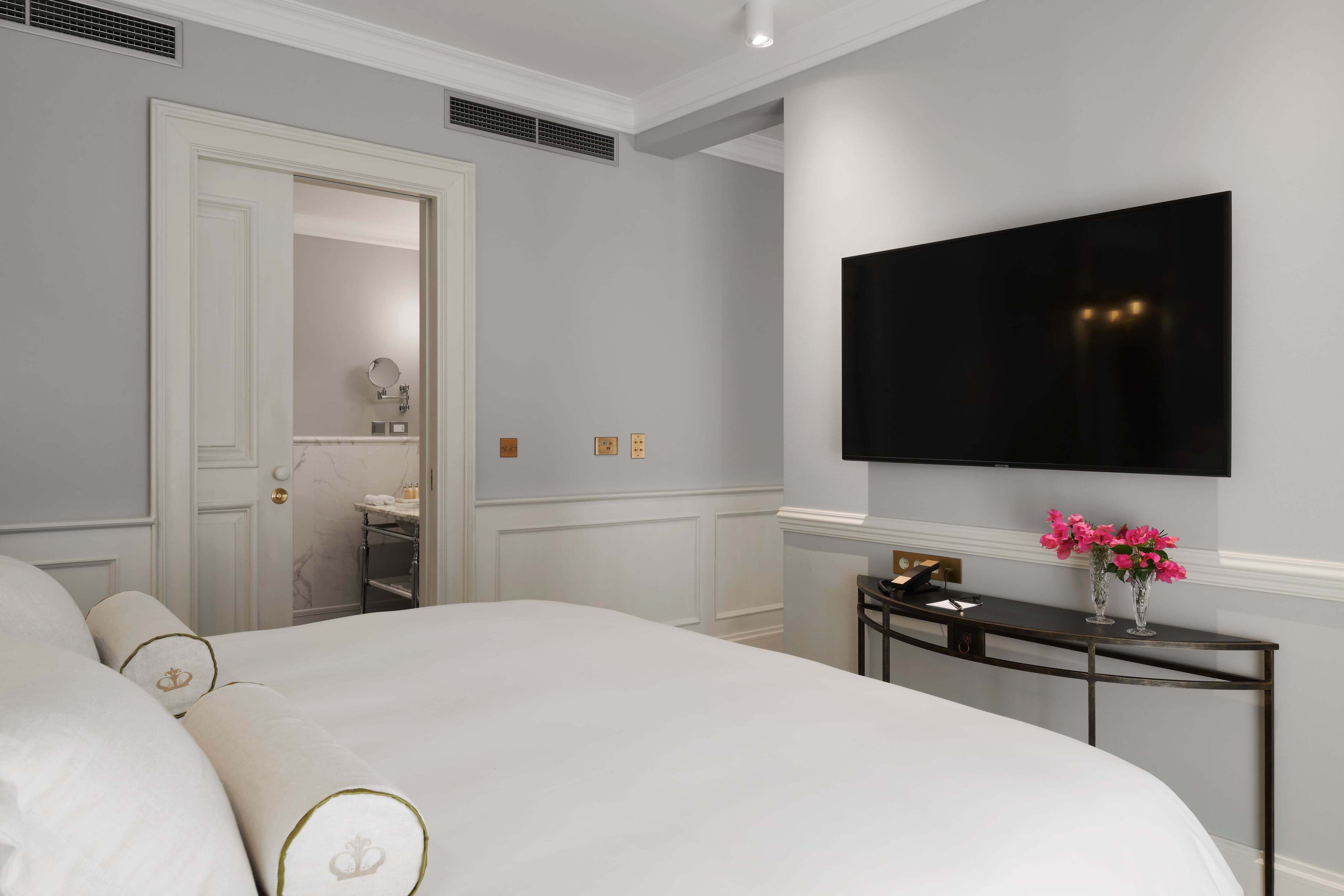A hotel room with a large bed, a flat-screen TV mounted on the wall, a small table with a vase of flowers, and a door leading to a bathroom.