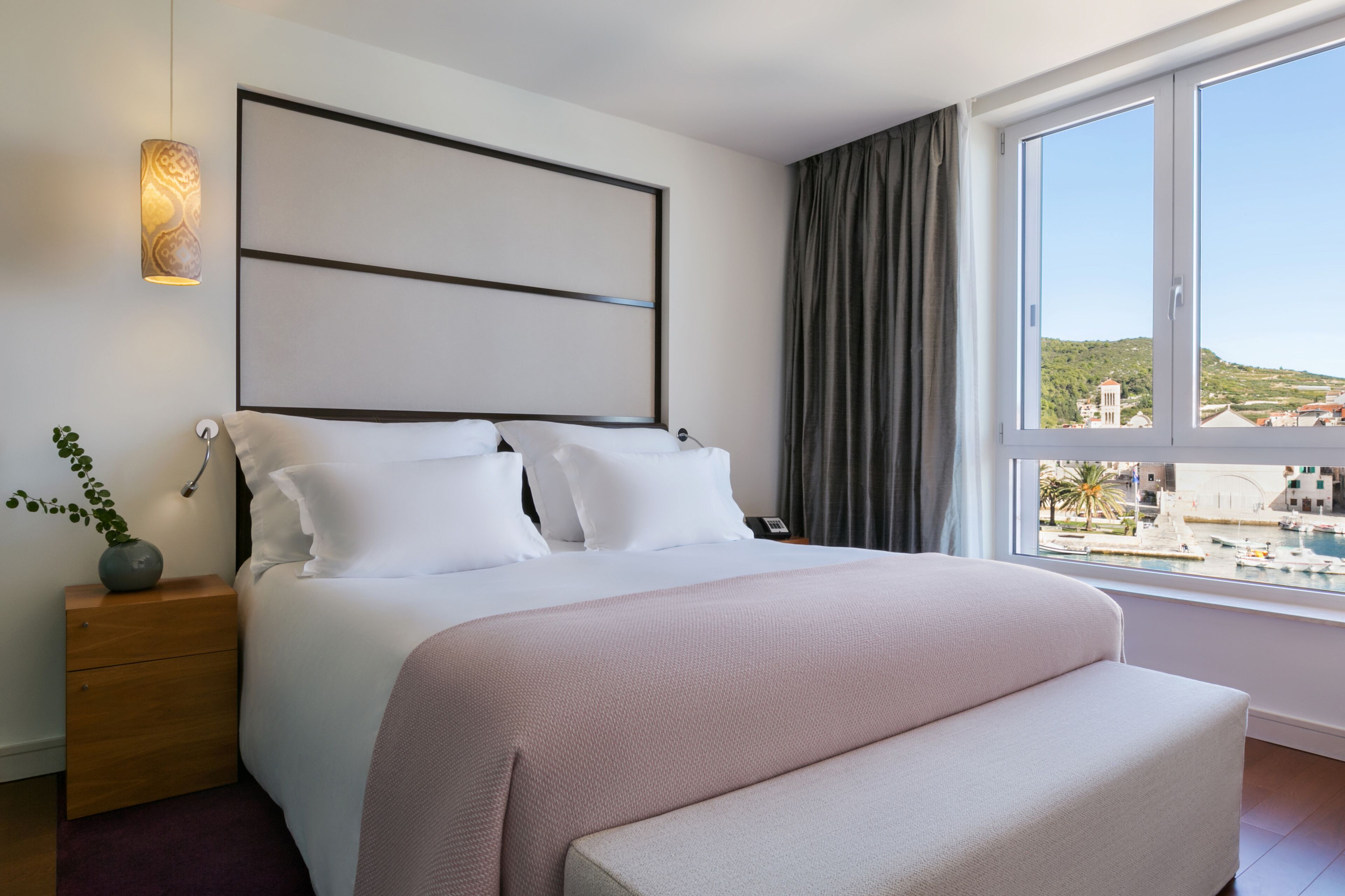 Ein Hotelzimmer mit einem großen Bett, einem Nachttisch mit Lampe, Blick auf einen Yachthafen und einer Stadtansicht im Hintergrund.