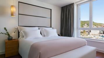 Ein Hotelzimmer mit einem großen Bett, einem Nachttisch mit Lampe, Blick auf einen Yachthafen und einer Stadtansicht im Hintergrund.