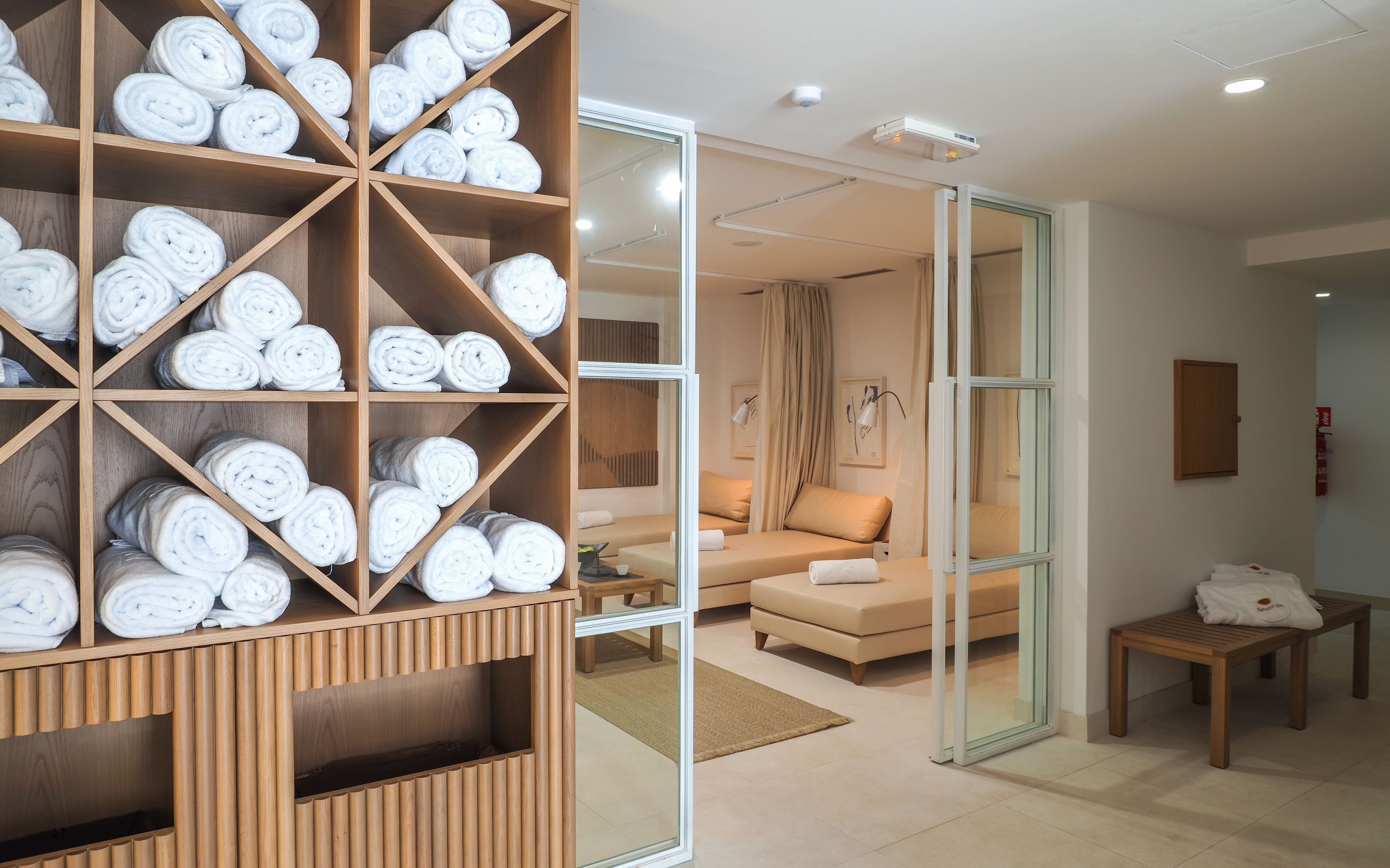 A hotel room with a wooden shelf holding rolled towels, a sofa, and a small table with a towel.
