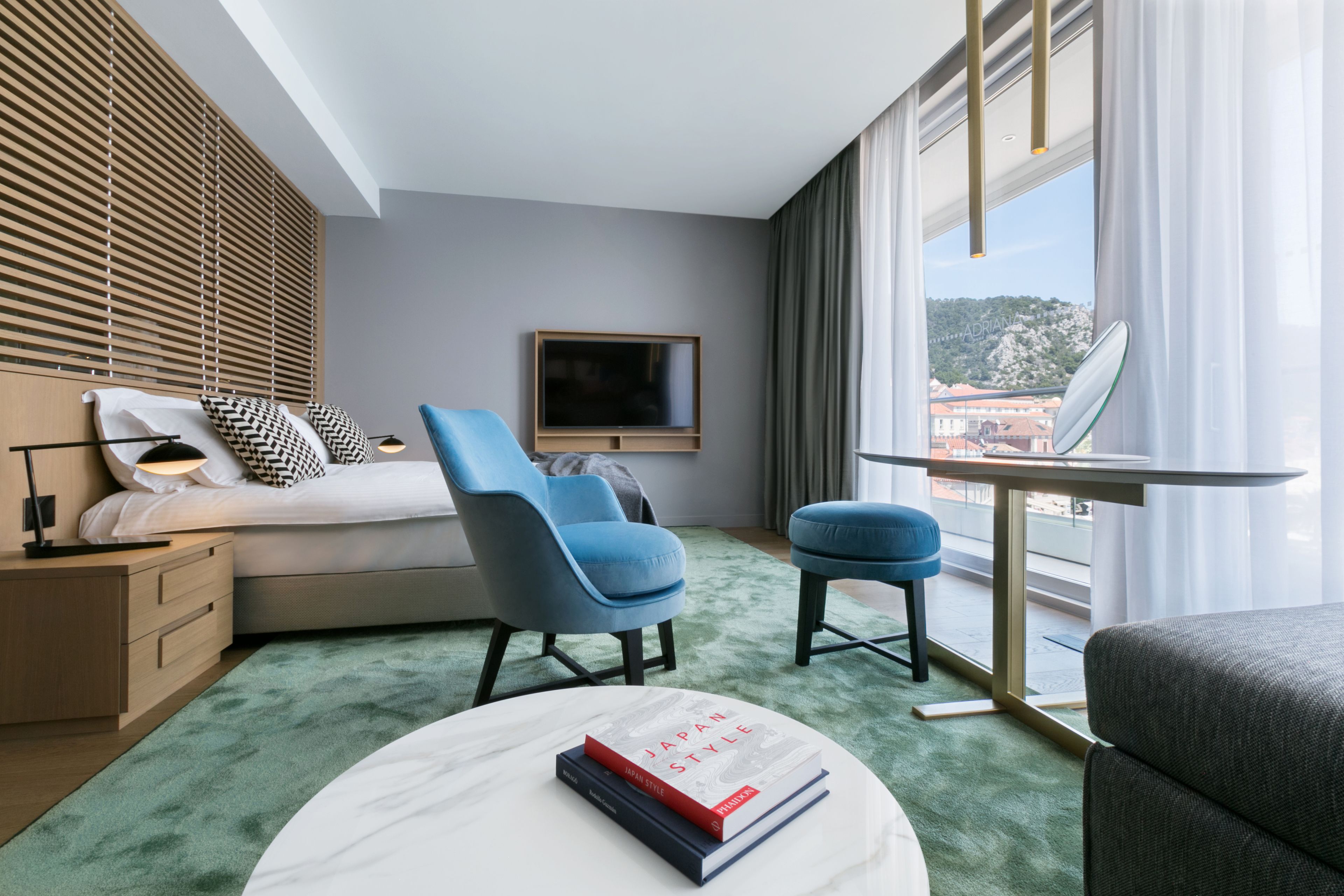 Une chambre d’hôtel moderne avec un lit, deux chaises bleues, une petite table avec des livres, un téléviseur et une vue sur le paysage urbain à travers de grandes fenêtres.