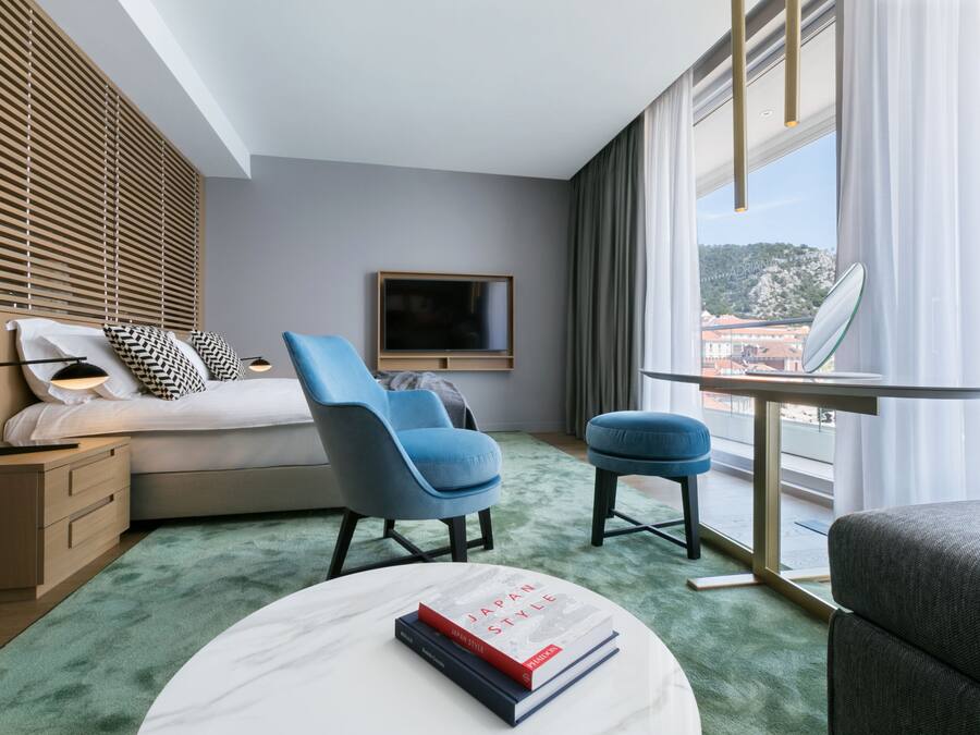 Une chambre d’hôtel moderne avec un lit, deux chaises bleues, une petite table avec des livres, un téléviseur et une vue sur le paysage urbain à travers de grandes fenêtres.