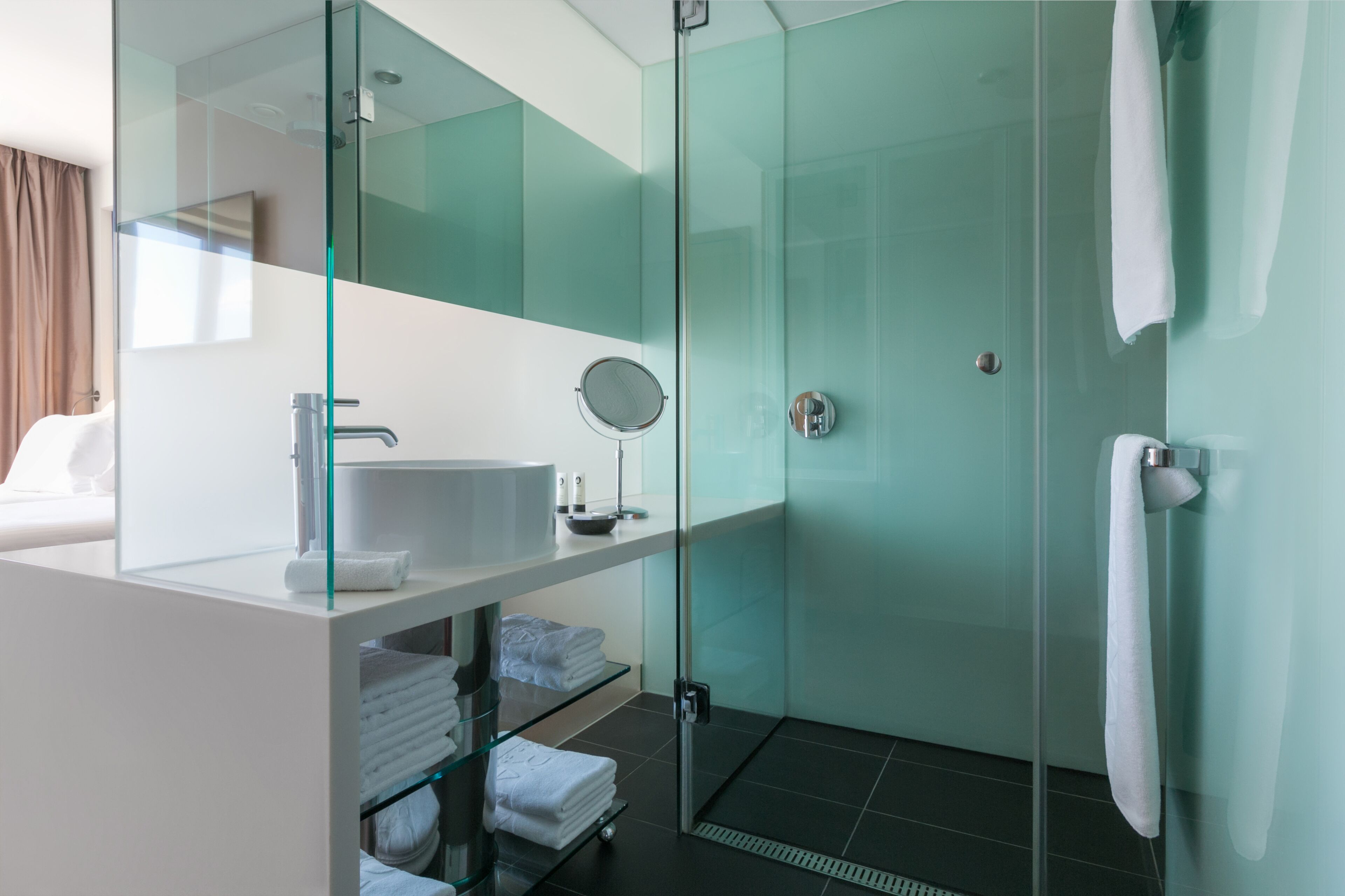 A modern bathroom with a glass shower enclosure, a white sink, and a glass shelf with folded towels.
