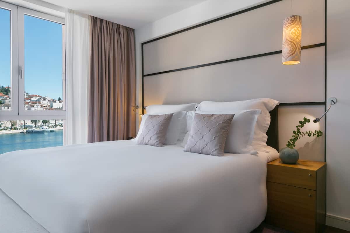 Ein Hotelzimmer mit einem großen Bett, einem Nachttisch, einer Pflanze in einer Vase und Blick auf das Meer und Gebäude.