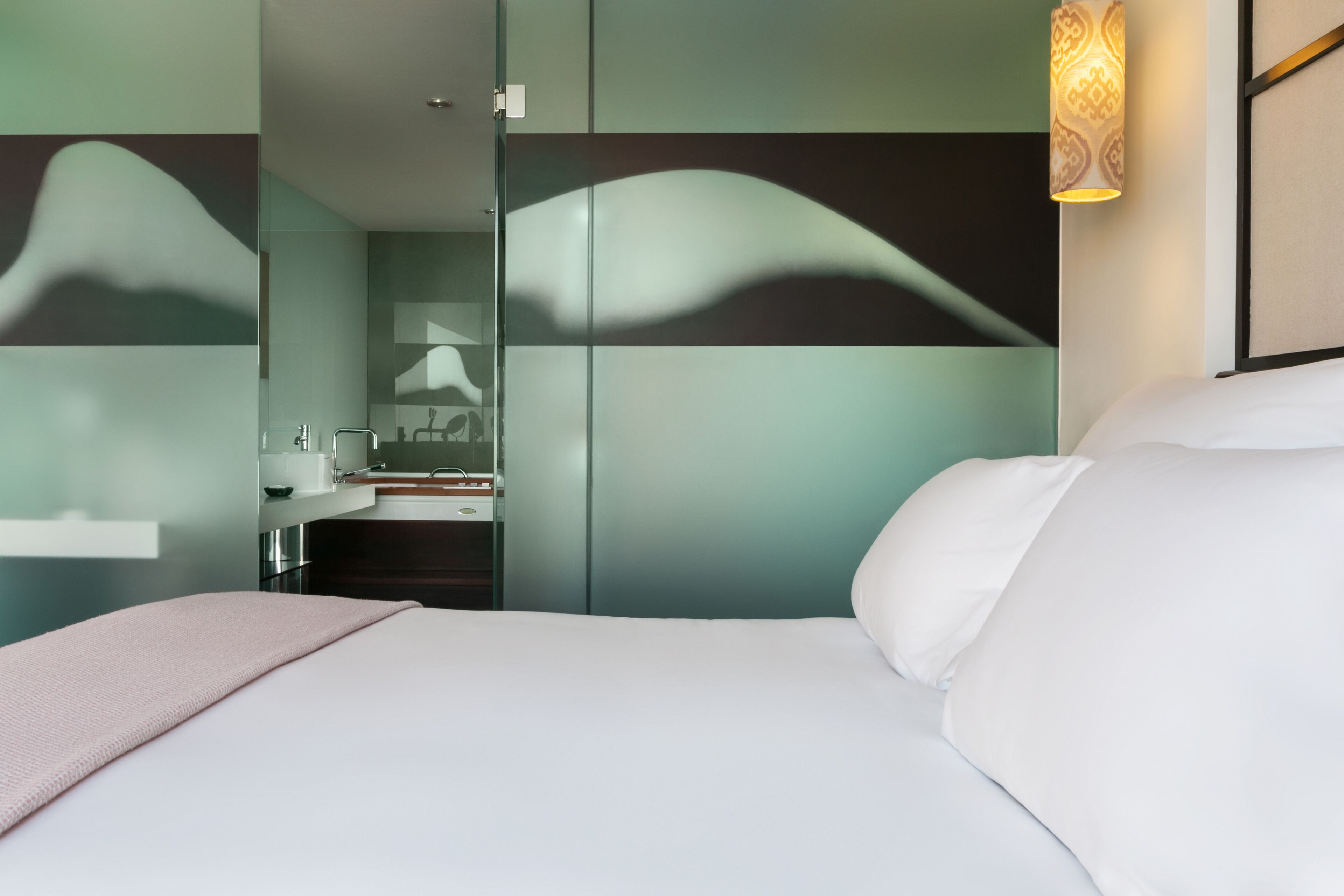 A modern hotel room with a bed, a bathroom with a mirror, and a wall-mounted light fixture.
