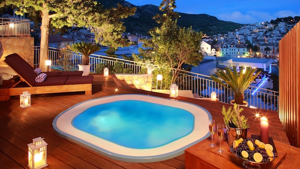 A wooden deck with a hot tub, lounge chairs, and a view of a hillside town at dusk. - Adriana Hvar Spa Hotel (Hvar)