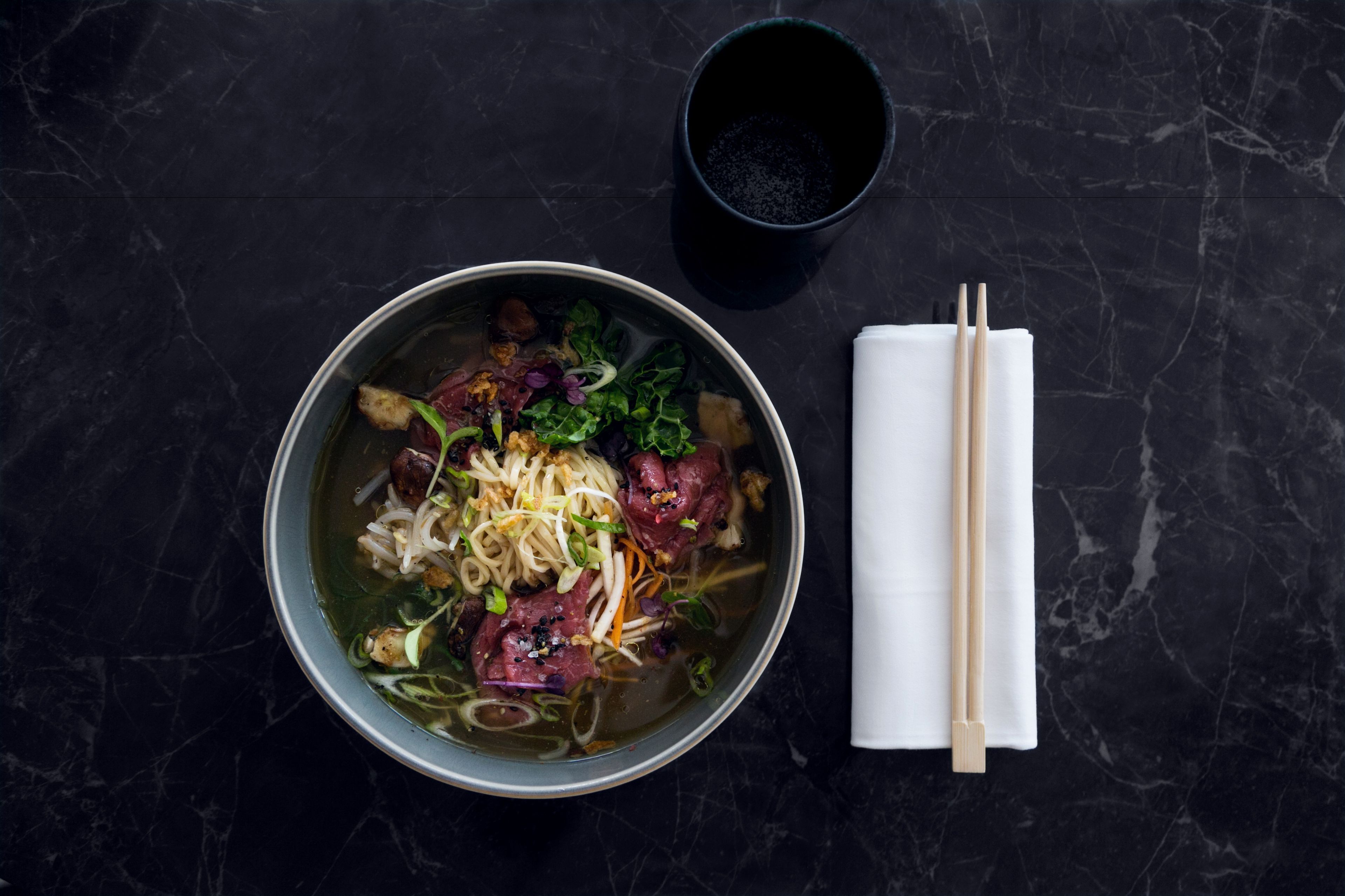 A bowl of noodle soup with meat and vegetables on a dark marble surface.