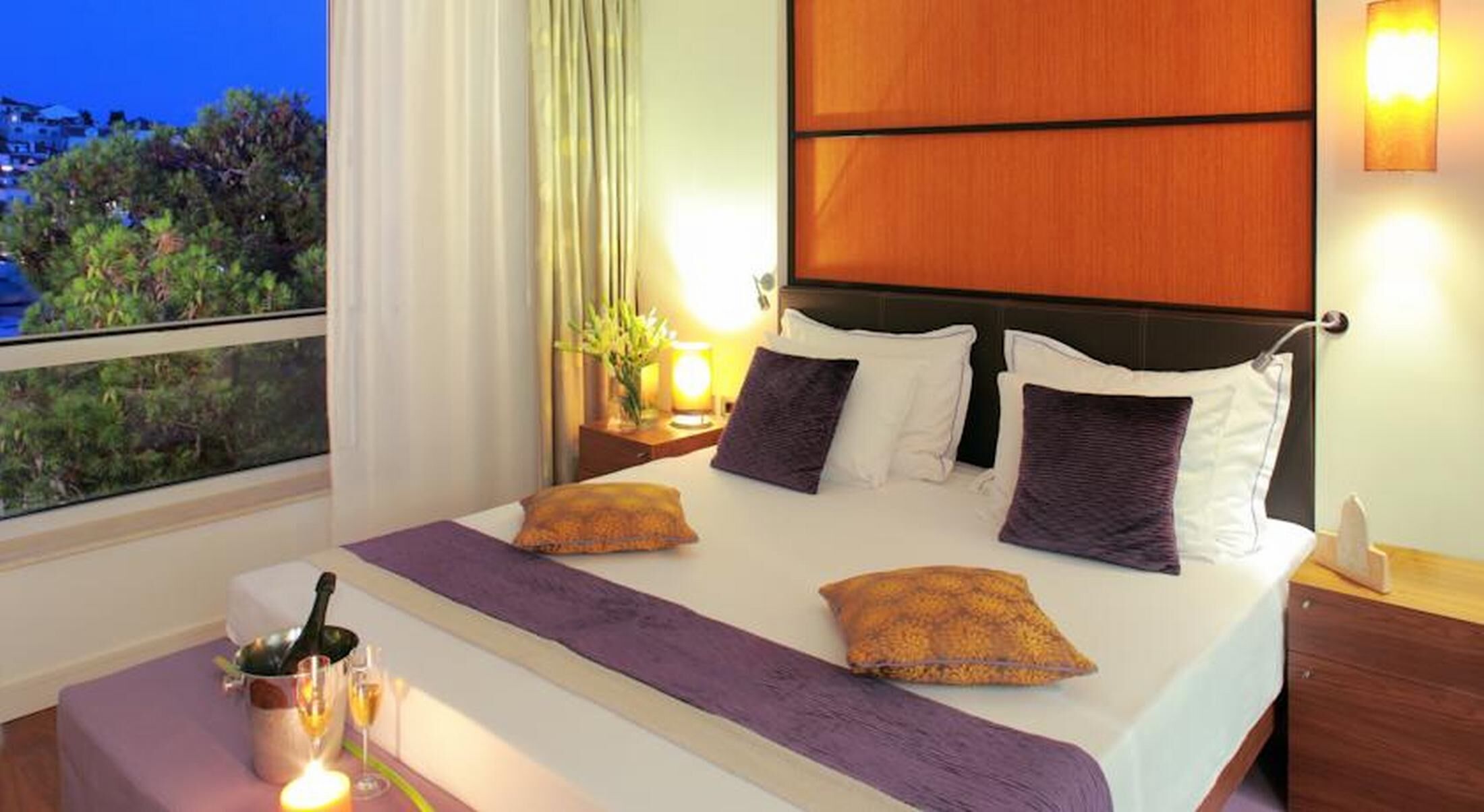 Habitación de hotel con cama, mesita de noche, ventana con vistas y una mesilla con una vela.