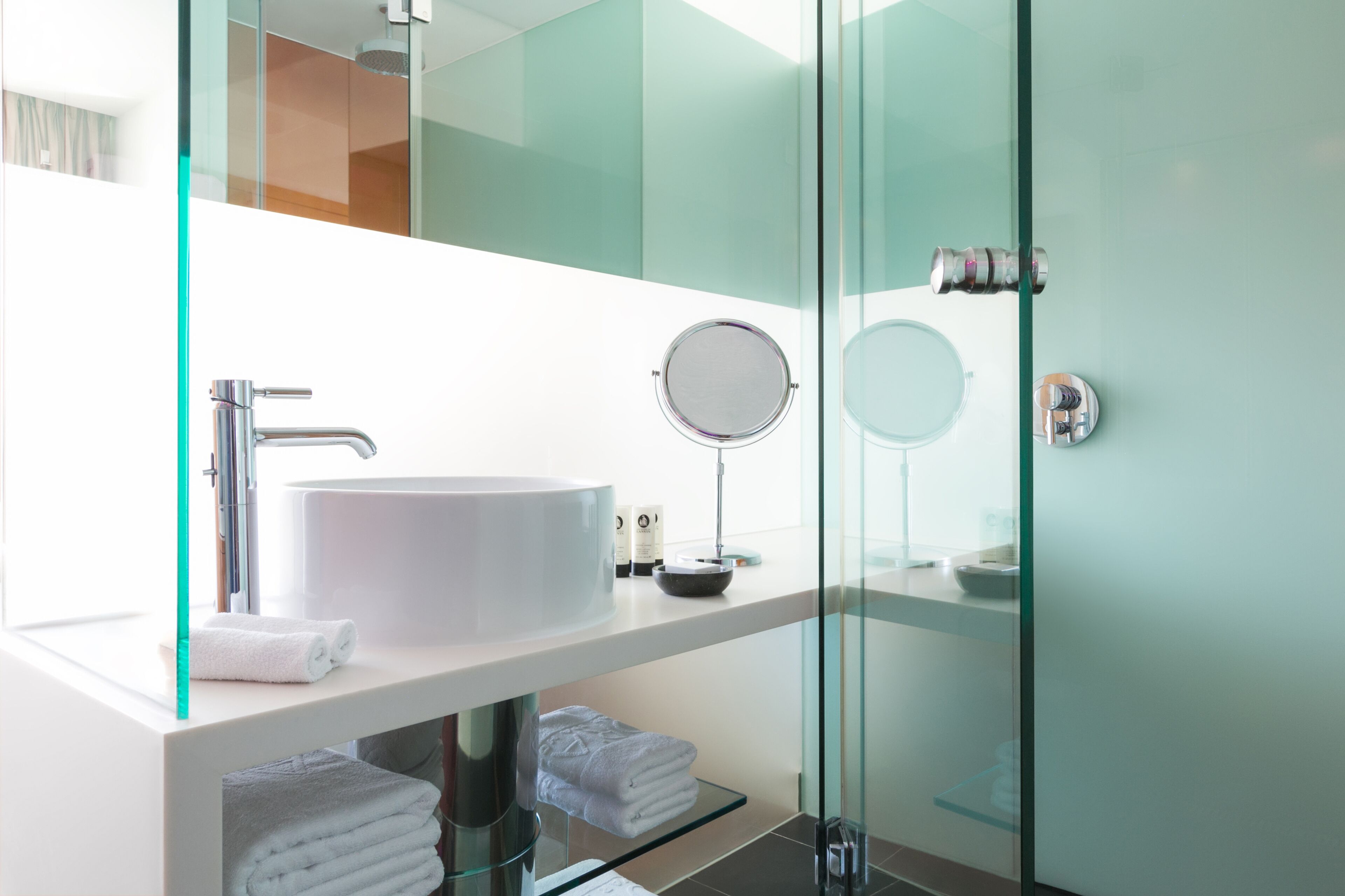Un baño moderno con un lavamanos grande de color blanco, un espejo redondo y cabina de ducha de vidrio.
