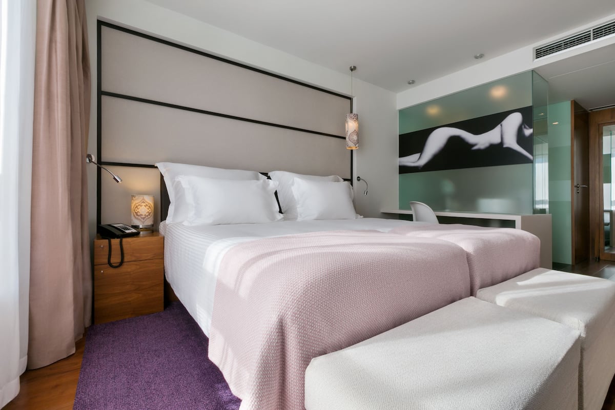 Ein modernes Hotelzimmer mit einem großen Bett, einem Nachttisch, einer Lampe und einer Glaswand mit Kunstwerken.