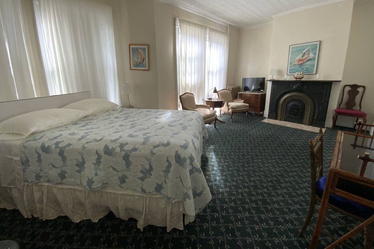 A hotel room with a large bed, a desk with a chair, a television, a fireplace, and two paintings on the walls.
