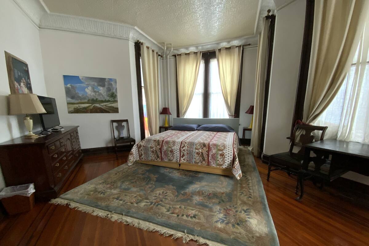 A bedroom with a large bed, wooden floor, a dresser, a desk, and a chair.